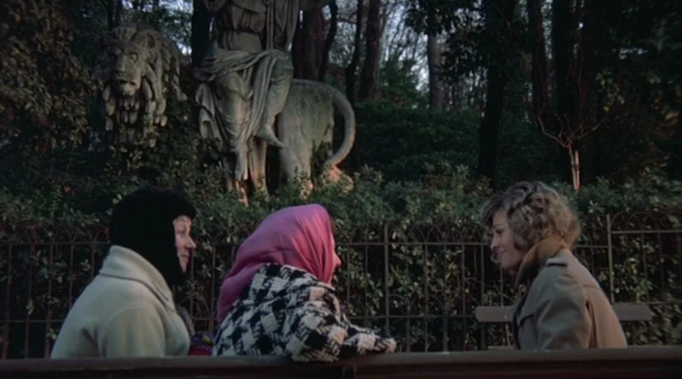 Don’t Look Now (1973) movie still: medium — A public park features a wrought-iron fence and a large stone lion statue background. Three women sit…