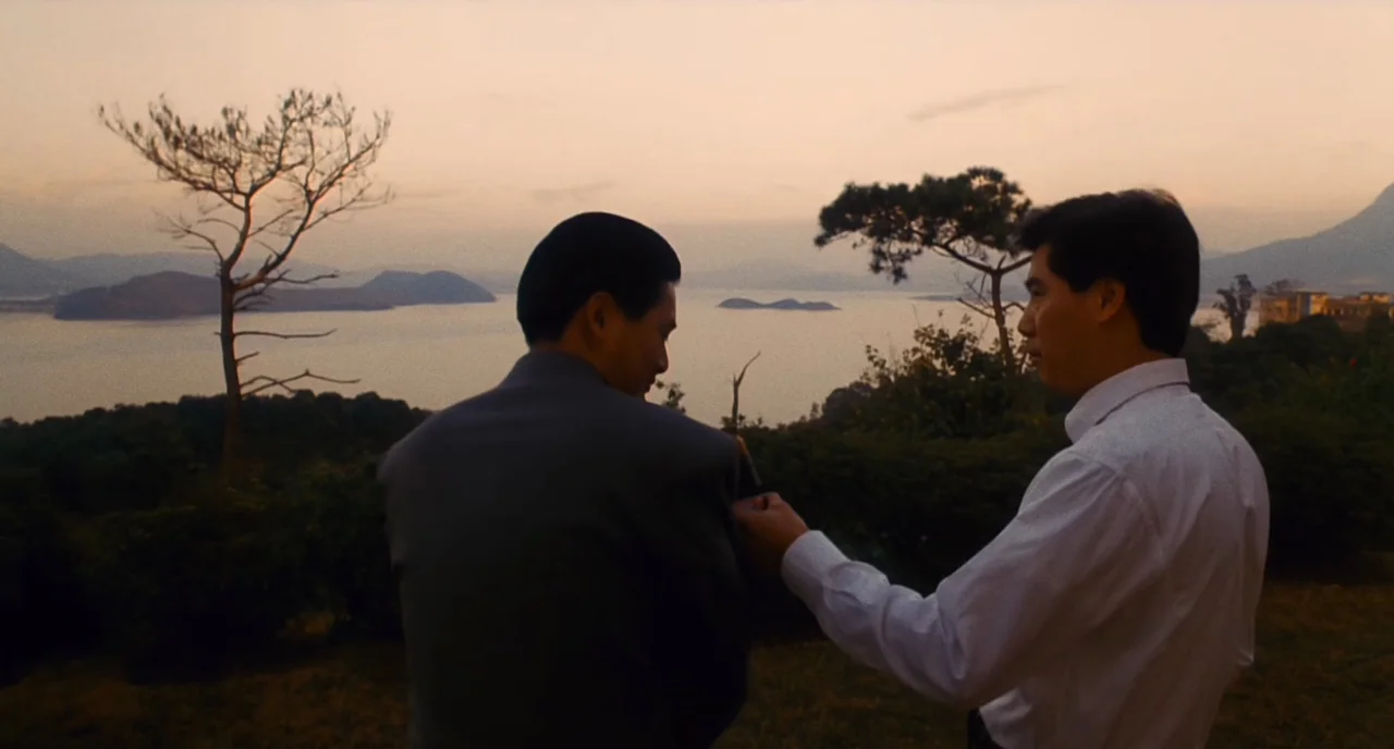 The Killer (1989) movie still: medium — Two men stand on a hillside overlooking a large lake during golden hour. A man adjusts the shoulder of a…