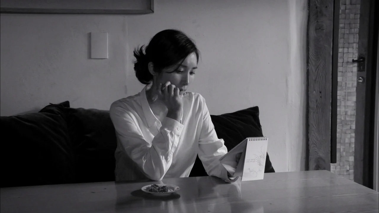 The Day He Arrives (2018) movie still: medium — A woman sits at a wooden table in a room with a plain wall. She holds a small sketchbook and looks…
