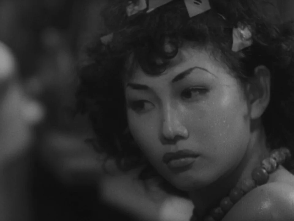 Stray Dog (1949) movie still: closeup — A close portrait in a dark interior space with soft lighting on the subject. The woman with curly hair looks…