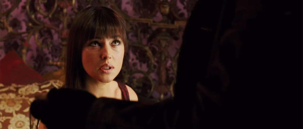 Shoot 'Em Up (2007) movie still: closeup — A dark bedroom features a wall with ornate purple and gold damask wallpaper. A woman looks up toward a…