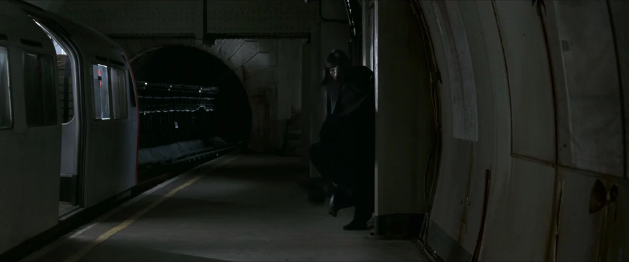 V for Vendetta (2006) movie still: wide — A subterranean train station platform features curved tiled walls and dark tunnel entrances. A person in…