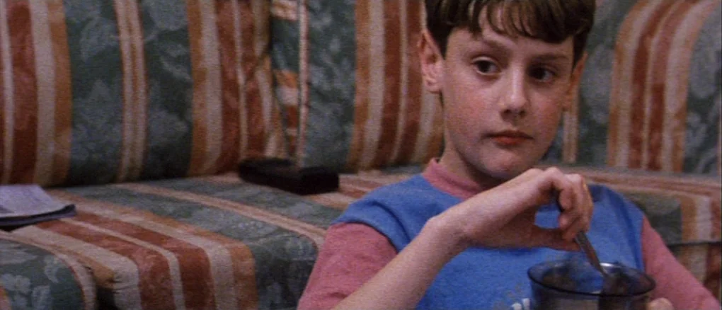 Wonderland (1999) movie still: medium — A young boy sits on a patterned green and orange couch inside a house. He holds a metal spoon and stirs the…