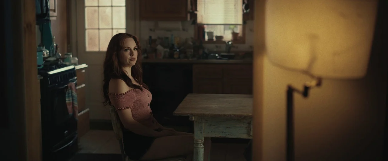 LaRoy, Texas (2023) movie still: medium — A dimly lit room with wood cabinetry and a window in the rear. A woman with red hair sits at a weathered…