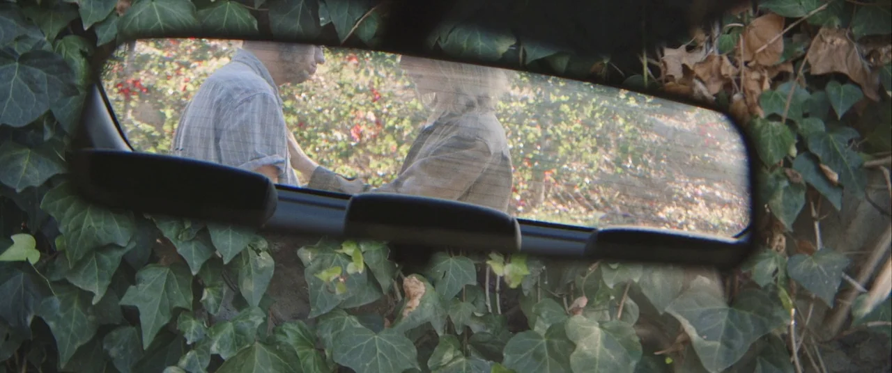 Always Shine (2016) movie still: closeup — An ivy-covered rearview mirror frames a reflection of two people standing outdoors. A man and a woman…