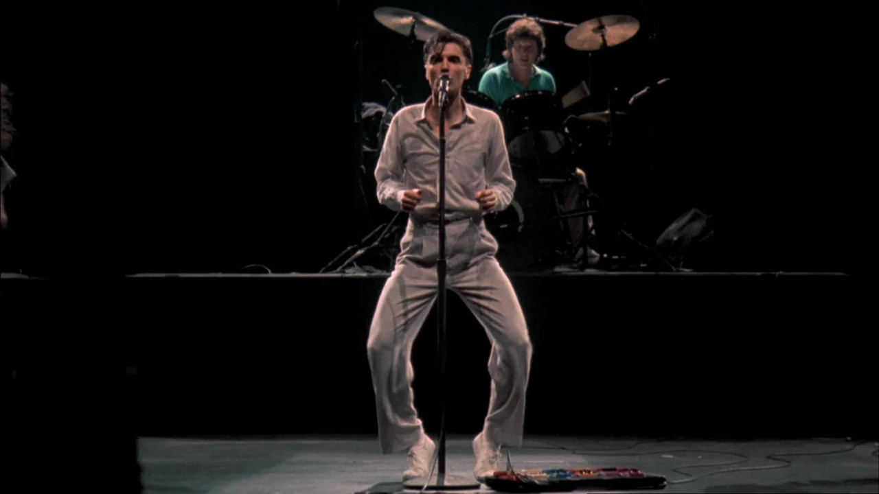 Stop Making Sense (1984) movie still: fullbody — A dark concert stage with a performer standing at a central microphone stand. A drummer is visible…