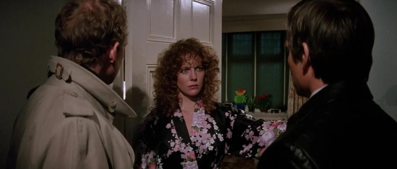 Lifeforce (1985) movie still: medium — A dark, residential doorway frames a woman standing between two men in coats. The woman in the floral robe…