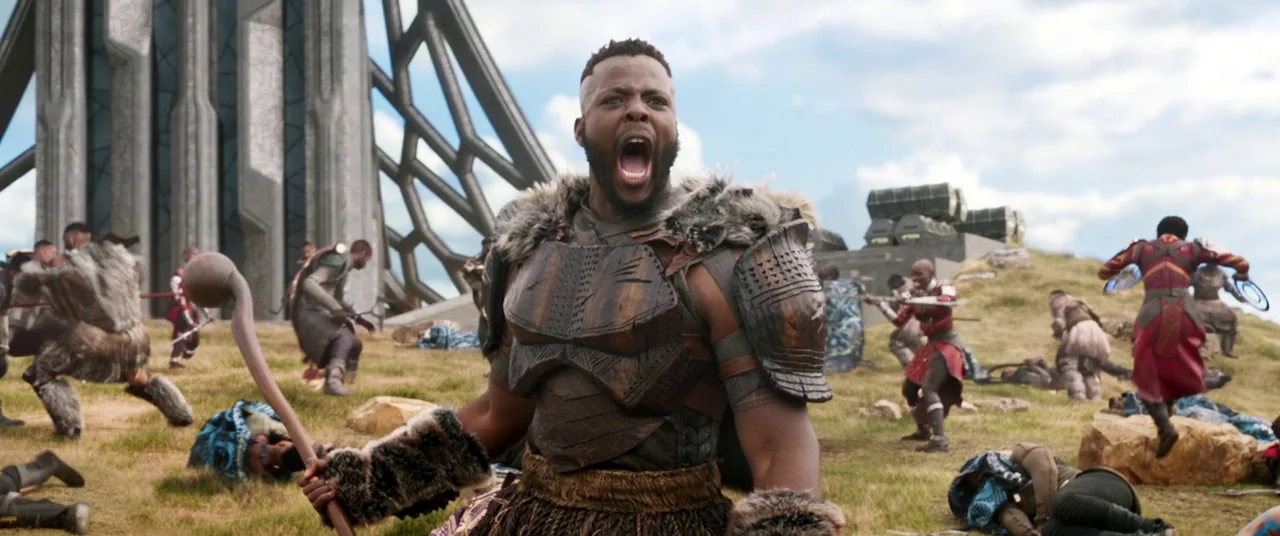 Black Panther (2018) movie still: medium — A grassy field with a towering metallic futuristic structure in the background. A man in tribal armor…