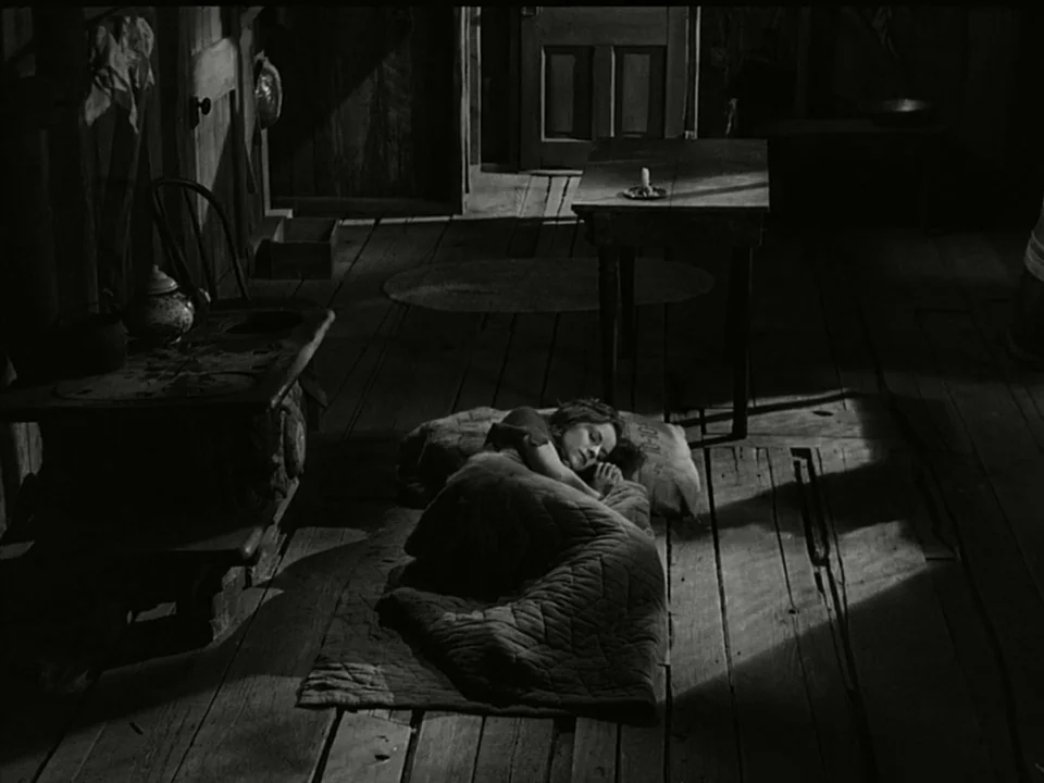 Modern Times (1936) movie still: wide — A rustic cabin interior with rough wooden floorboards and a lone table. A young woman lies asleep on a thin…