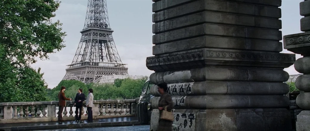 Munich (2005) movie still: wide — A wide stone bridge features thick masonry pillars adorned with scattered posters and flyers. Three men stand…