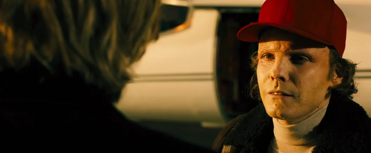 Rush (2013) movie still: medium — An airport tarmac during the golden hour with a white aircraft visible in the background. A man in a red cap…