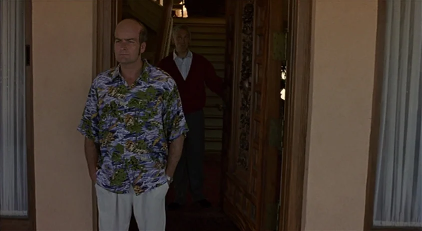 Being John Malkovich (1999) movie still: fullbody — A man in a patterned shirt stands on the concrete porch of a house. A second man in a maroon…