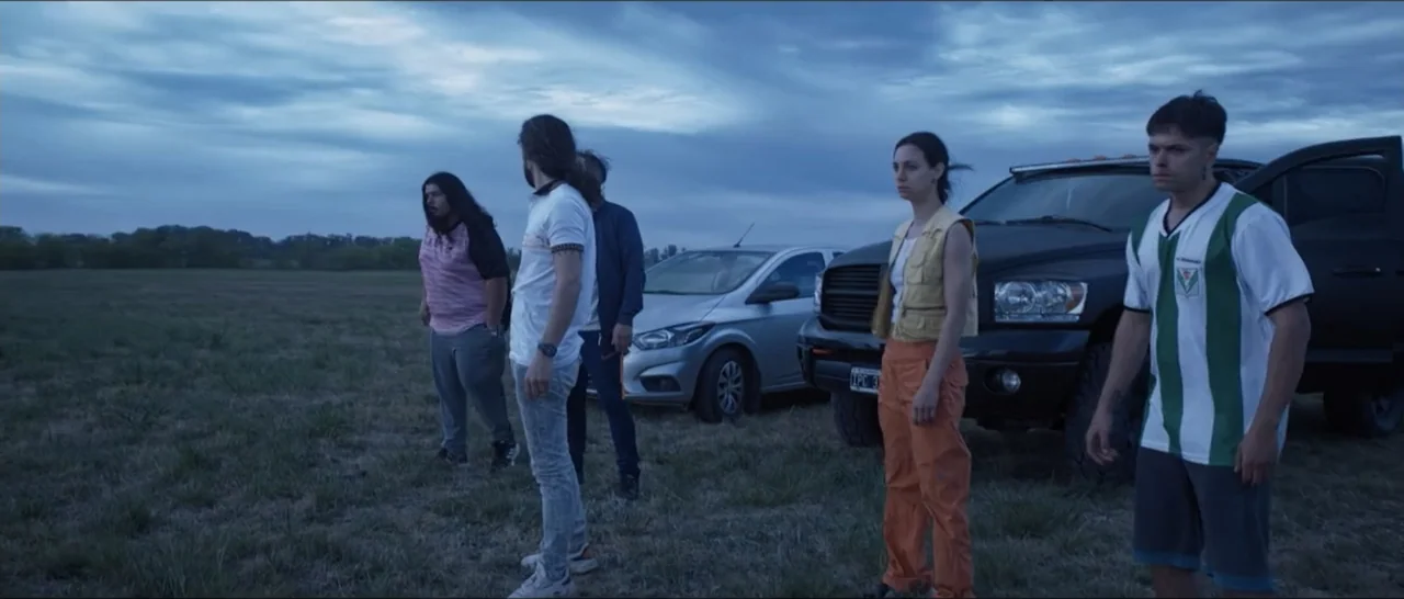 Gunman (2025) movie still: wide — An open rural field under a cloudy dusk sky features two stationary vehicles parked nearby. A group of five young…