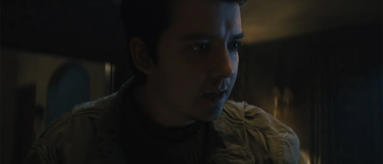 All Fun and Games (2023) movie still: closeup — A dimly lit interior space with shadows obscuring the surrounding furniture and walls. A young man…
