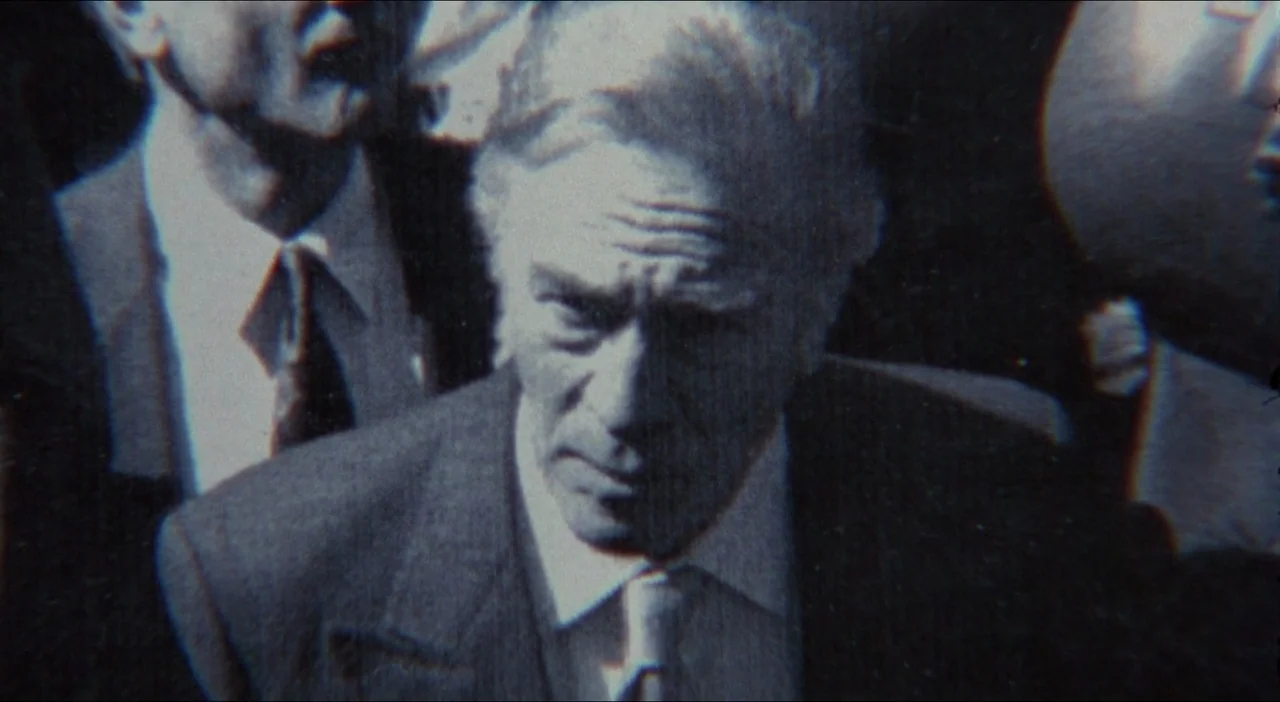 Twelve Monkeys (1995) movie still: closeup — An outdoor urban crowd surrounds an older man wearing a suit and tie. The image has a degraded, analog…