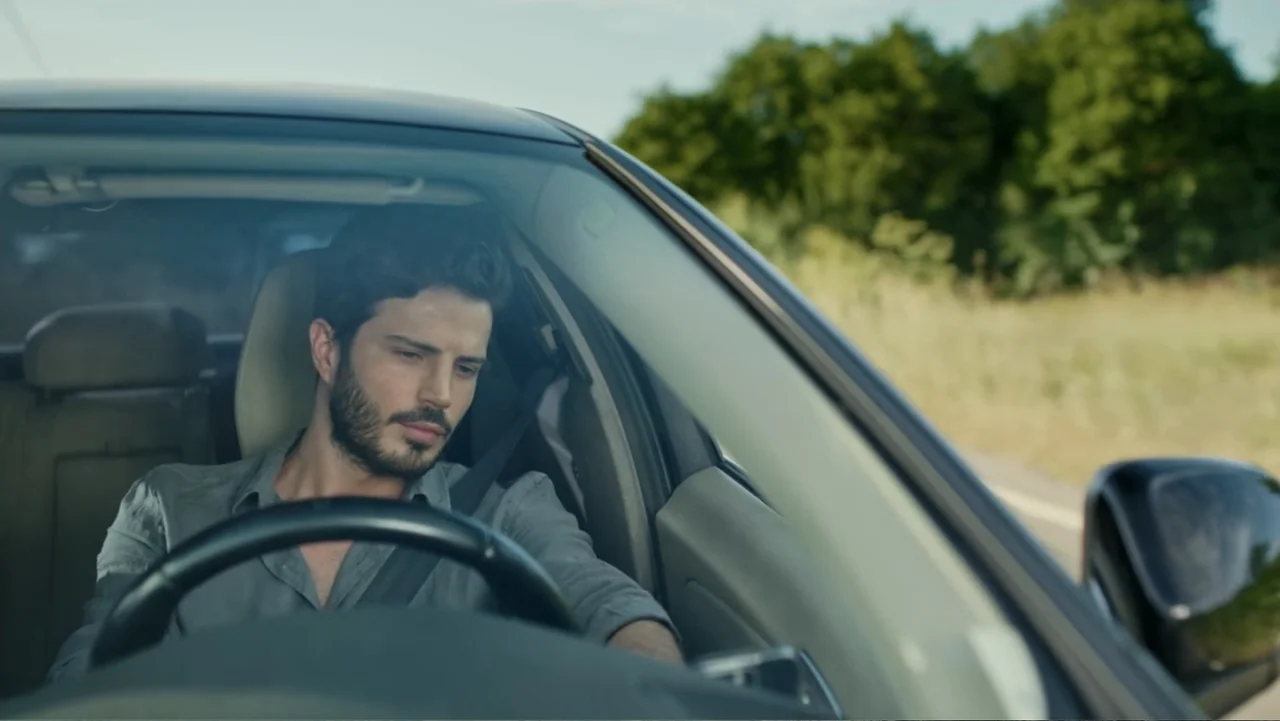 Paranormal Cuma (2024) movie still: medium — A man drives a car along a road lined with trees during the day. He sits behind the steering wheel…