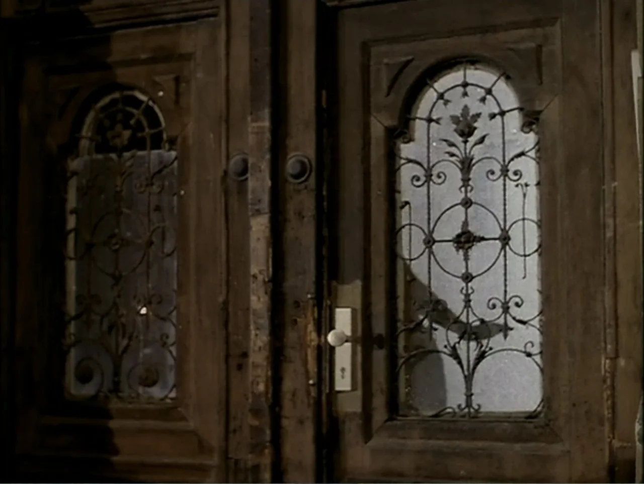 A Change of Place (1994) movie still: medium — A pair of weathered, dark-stained wooden double doors feature arched glass panes. Decorative black…