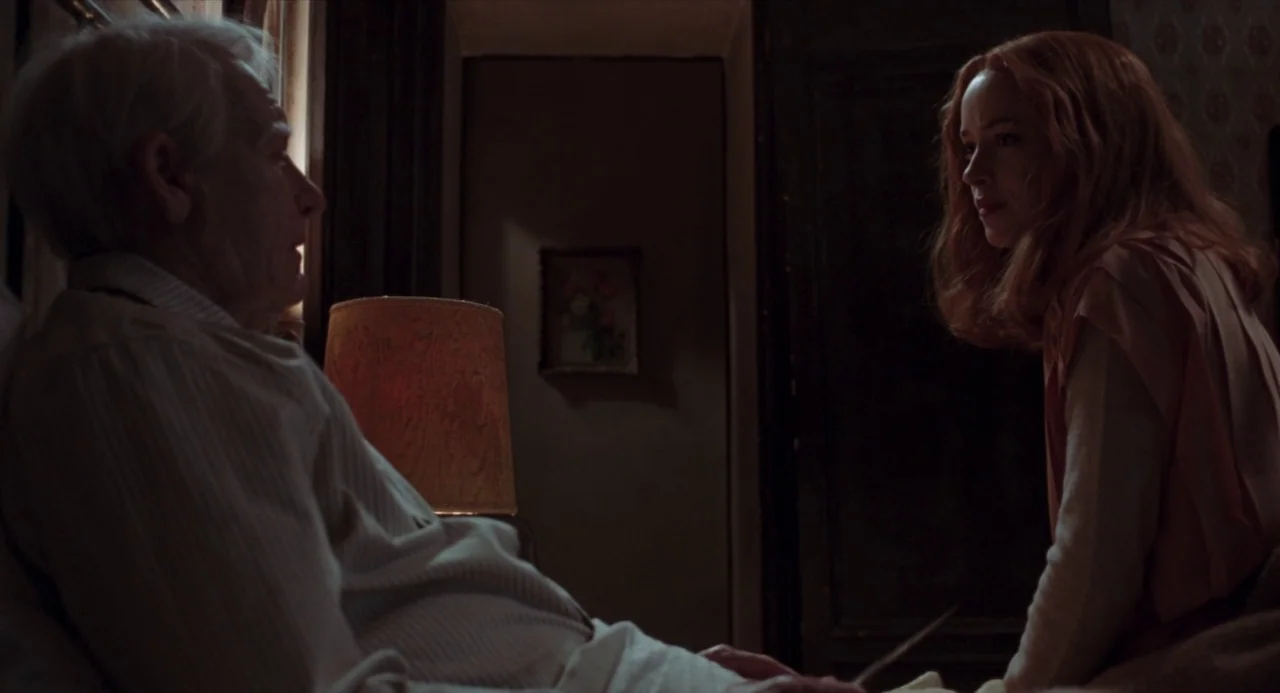 Suspiria (2018) movie still: medium — A dimly lit room with wallpaper features an elderly man and a young woman. The woman sits beside the bed while…