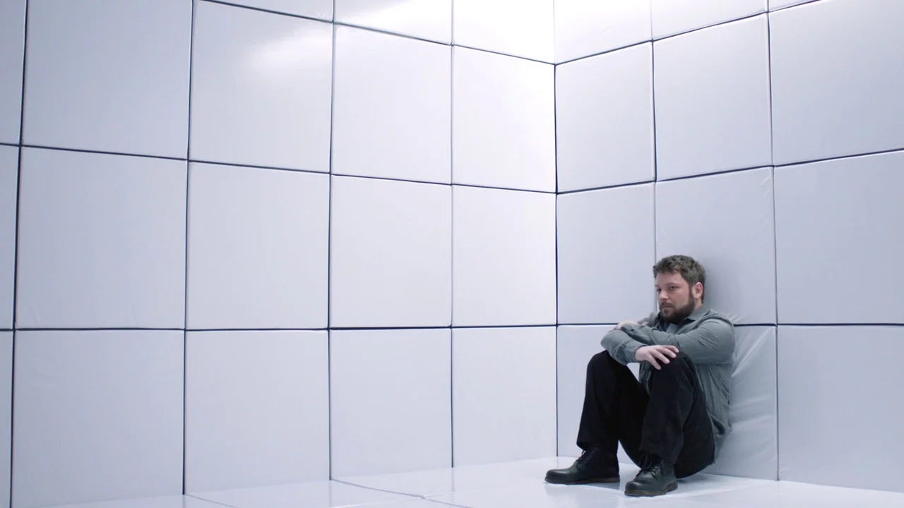 Cult of Chucky (2017) movie still: wide — A windowless room features walls and floor lined with white, square-tiled padded panels. A man in dark…