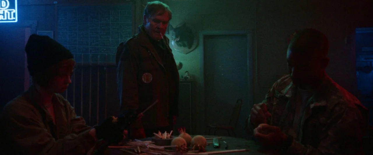 VFW movie still: medium — A dimly lit, sparse bar environment contains a dark table with wooden game pieces. Three individuals sit or stand around…