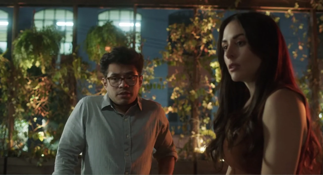 Casando a mi Ex (2023) movie still: medium — A dark balcony at night is filled with hanging plants against a windowed wall. A man and a woman stand…