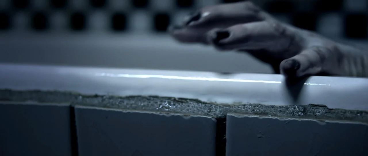 The Lullaby (2019) movie still: closeup — A close view of a white ceramic bathtub edge with chipped tile grout. A pale hand with dark fingernails…