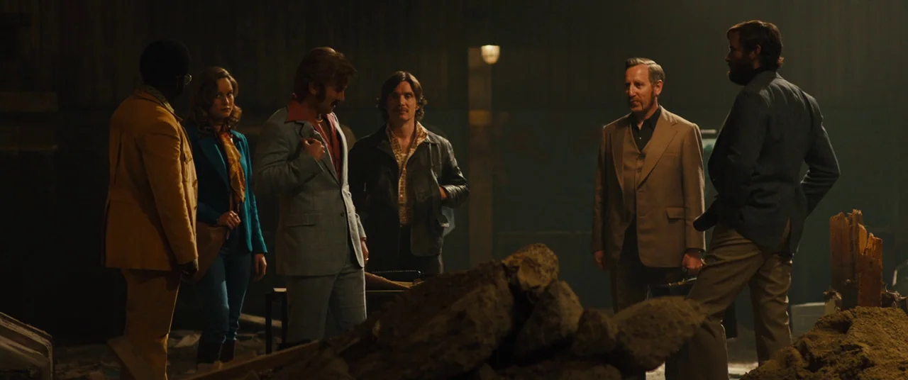 Free Fire (2016) movie still: wide — A dimly lit industrial environment filled with rubble and construction debris on the ground. Six people in…