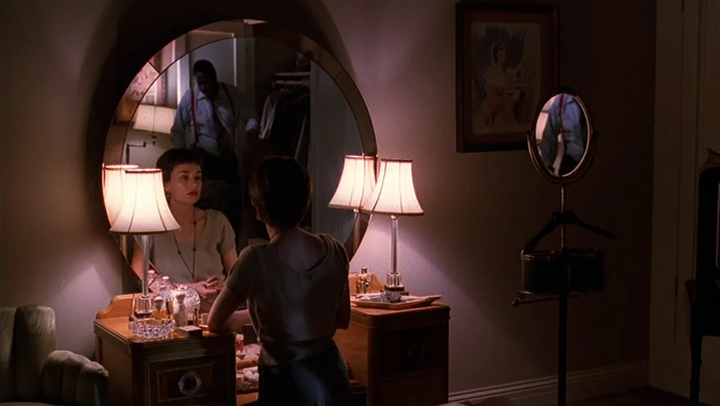 Bird (1988) movie still: medium — A dimly lit room contains a large round wooden vanity with twin table lamps. A woman faces her own reflection…