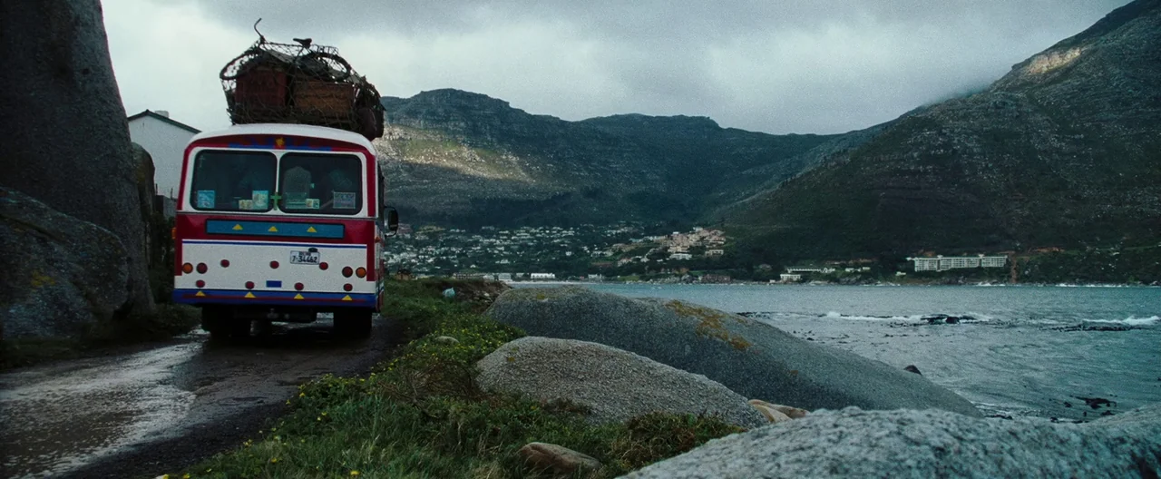 Lord of War (2005) movie still: wide — A rugged coastal road winds between large granite boulders and the ocean shoreline. A red and white bus…