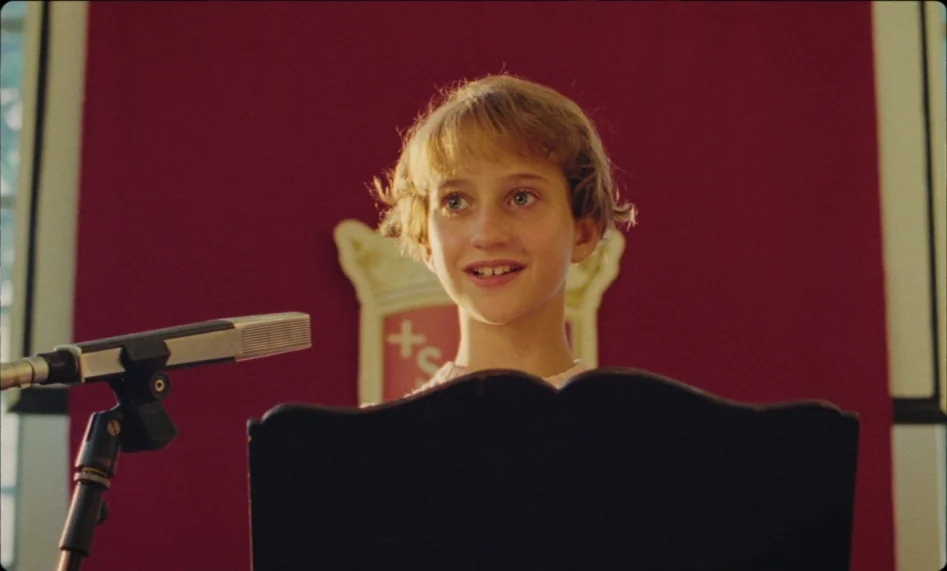 Misunderstood (2014) movie still: closeup — A child stands on a stage in front of a deep red banner. She speaks into a silver microphone placed on a…