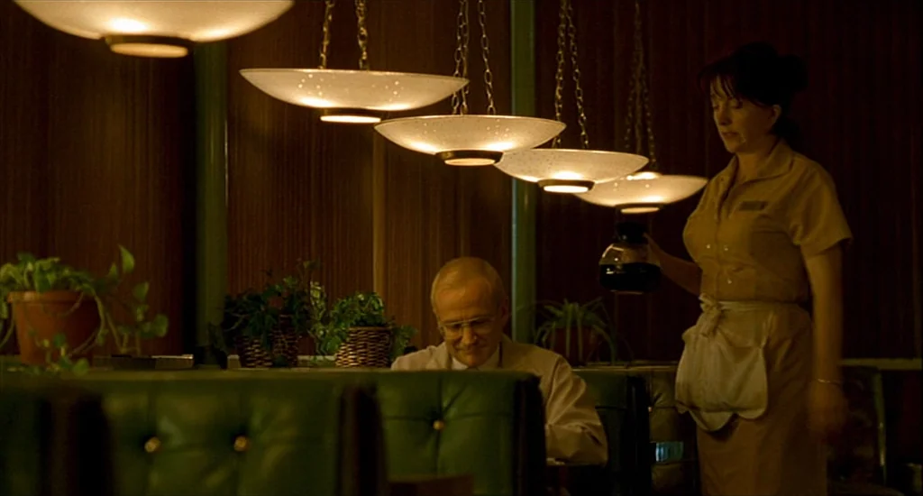 One Hour Photo (2002) movie still: medium — A 1990s diner features dark wood walls and green vinyl booths under pendant lights. A waitress in a tan…