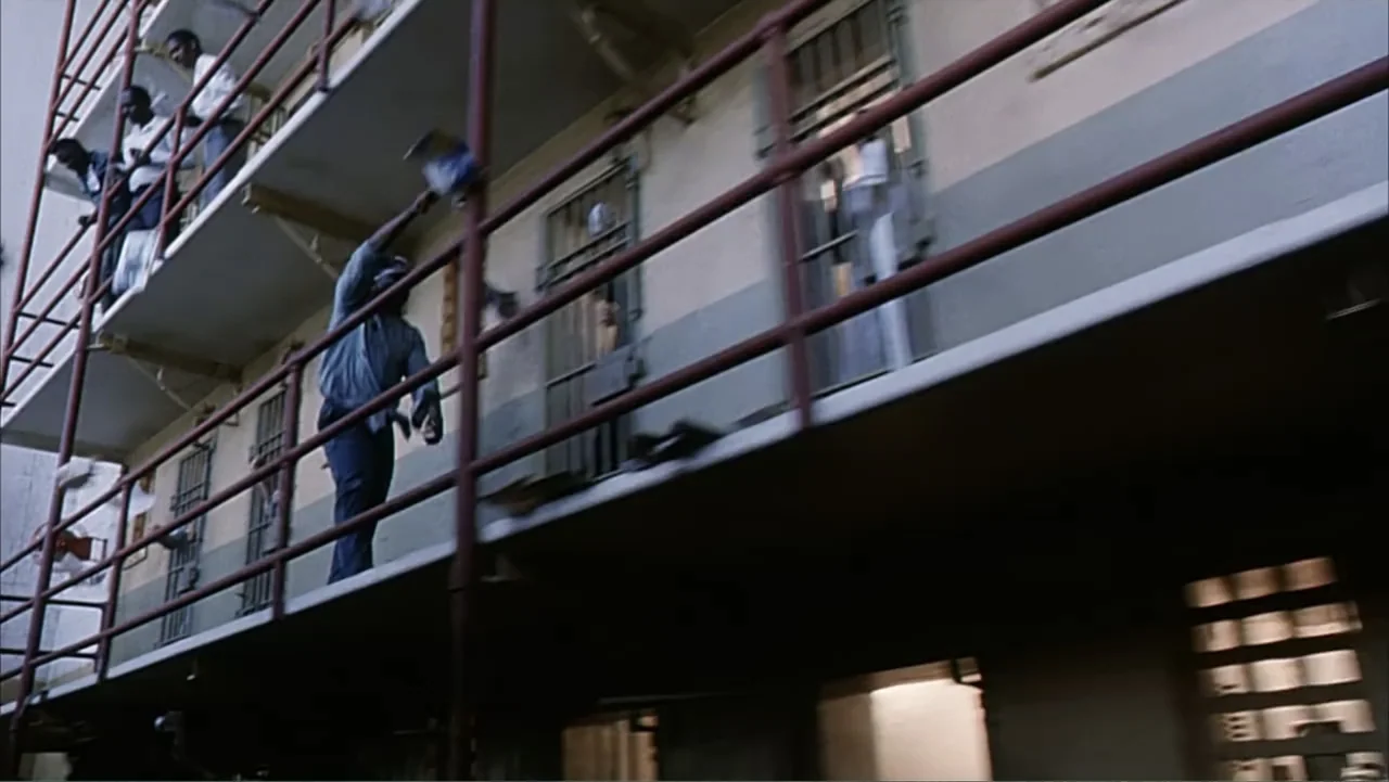 Against the Wall (1994) movie still: medium — A multi-level metal catwalk runs alongside a concrete building with barred doorways. A man stands on…
