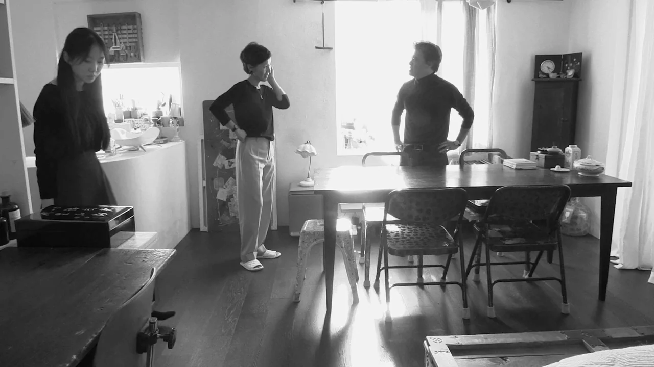 Walk Up (2022) movie still: wide — A sparse room features a dark wooden dining table and a large window. Three people stand within the space while…