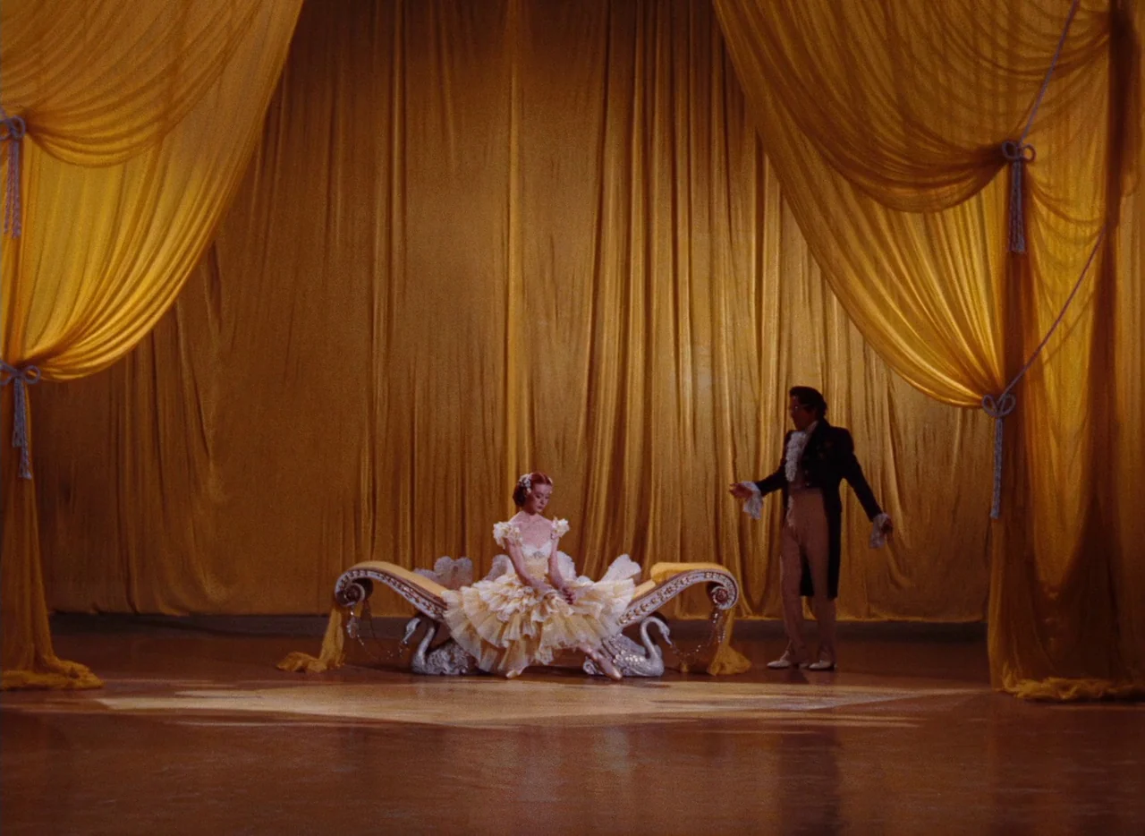 The Tales of Hoffmann (1951) movie still: wide — A theatrical stage features heavy, floor-to-ceiling yellow draped fabric backdrops. A woman in a…