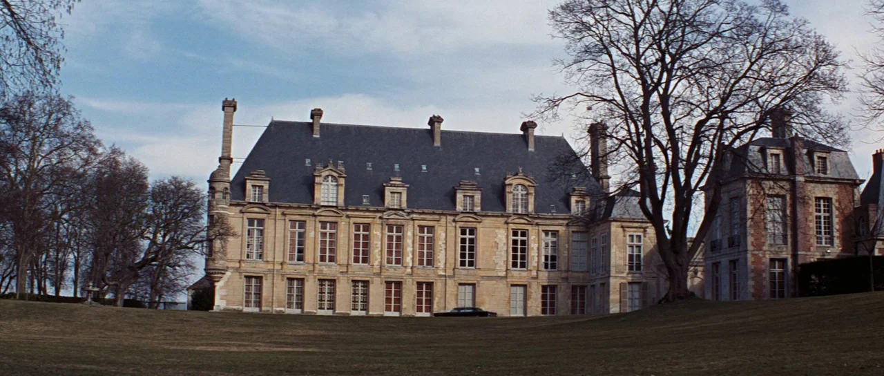 Thunderball (1965) movie still: establishing — A large stone chateau with a slate roof is situated on a grassy landscape. A dark vehicle is parked…