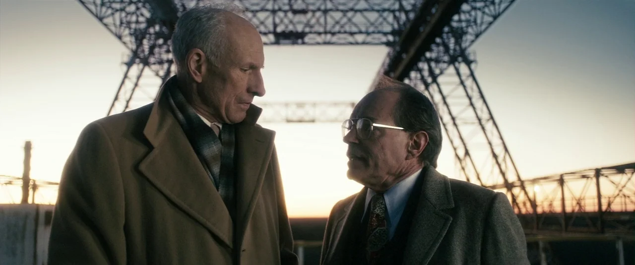 The Box (2009) movie still: medium — Two men stand on an elevated iron structure during a clear sunset. They face each other, positioned against the…