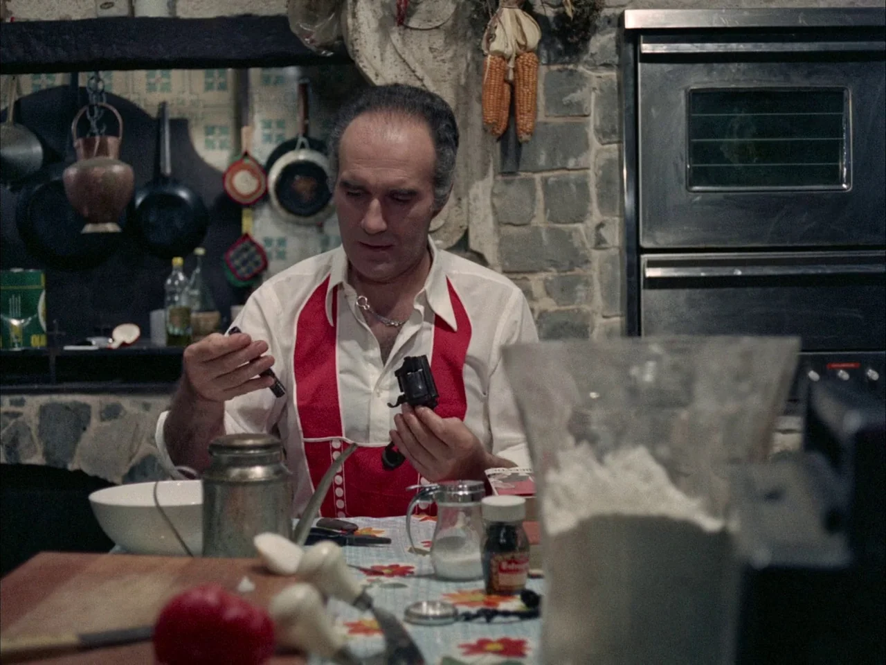 Dillinger is Dead (1969) movie still: medium — A rustic kitchen features stone walls adorned with dried corn and hanging cookware. A middle-aged man…