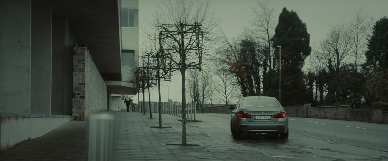 The Cellar (2022) movie still: wide — A paved sidewalk runs alongside a modern stone-walled building and a road. A person walks away into the…