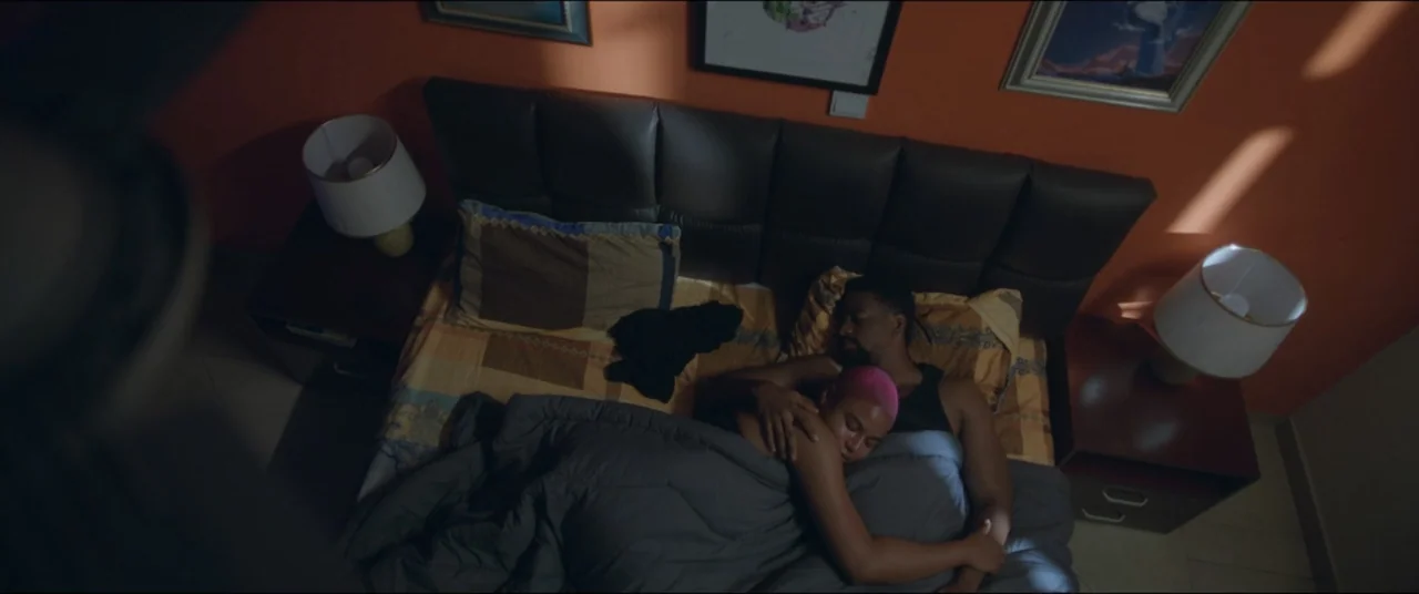 Saving Onome (2024) movie still: wide — A modern bedroom features orange walls and a leather headboard above a shared bed. A man and a woman lie in…