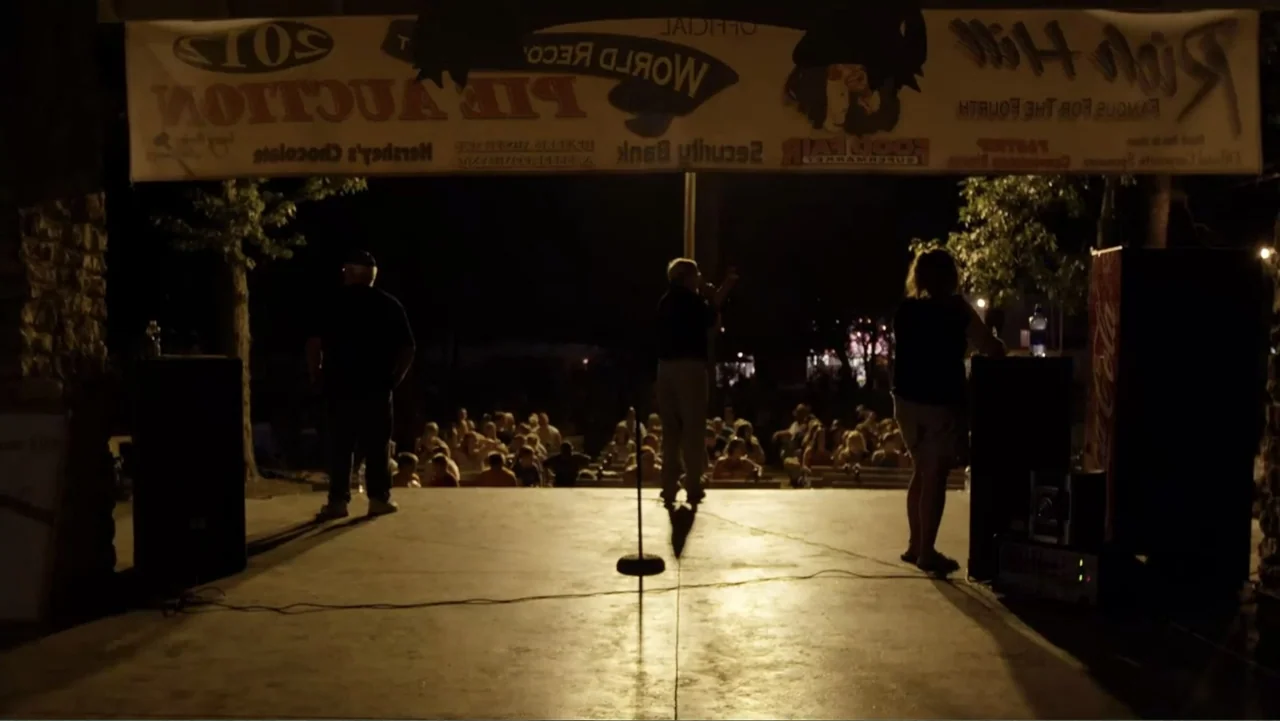 Rich Hill (2014) movie still: fullbody — An outdoor stage at night holds three people performing under bright overhead floodlights. The background…