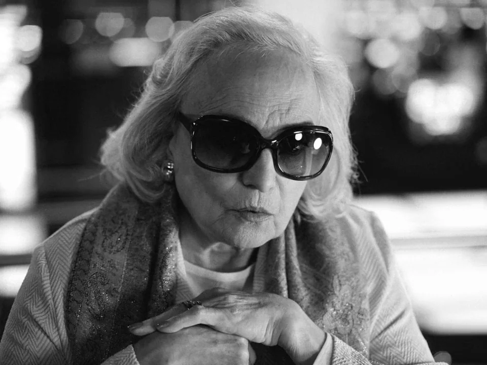 Tabu (2012) movie still: closeup — An older woman sits at a table in a dimly lit restaurant at night. She looks forward with her hands clasped,…