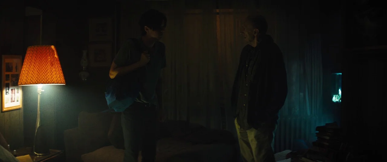 Daniel Isn’t Real movie still: fullbody — A dim living room with wood-paneled walls and a table lamp on a side table. An elderly man and a young man…