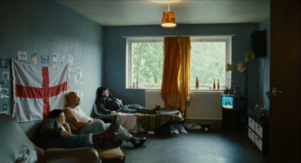 This Is England (2006) movie still: wide — A sparse, blue-walled room contains a flag of England pinned to the wall above a couch. Two people sit in…