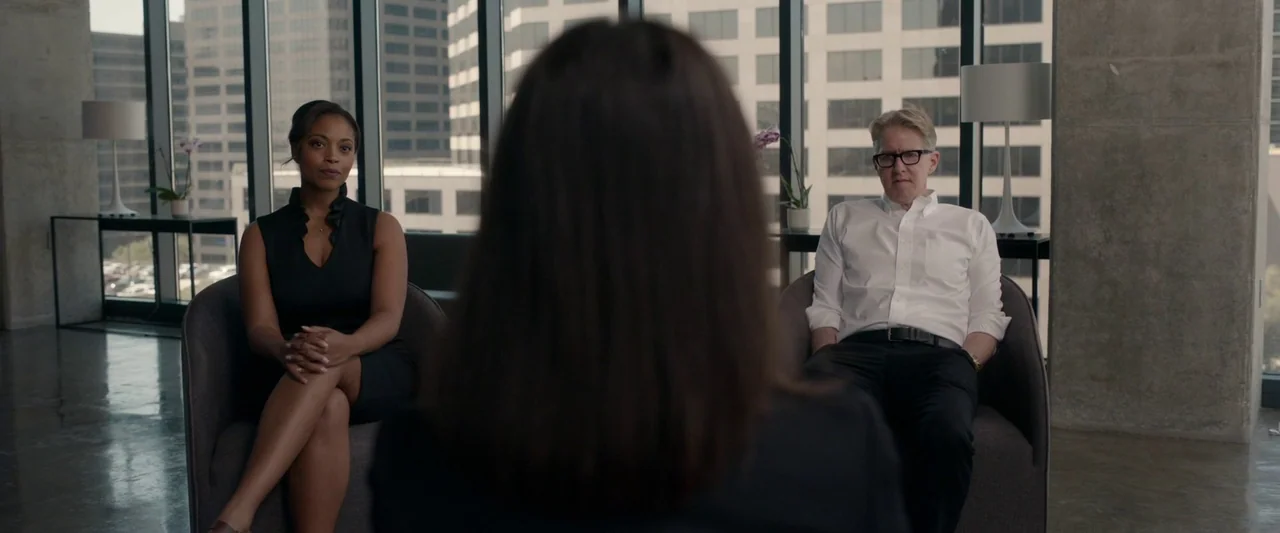 The Hunt (2020) movie still: medium — A high-rise meeting room with floor-to-ceiling windows overlooking a city business district. A man and a woman…