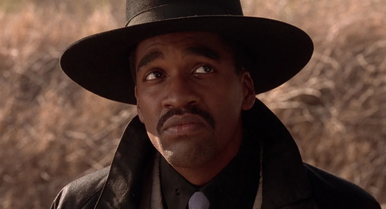 Tales from the Crypt: Demon Knight (1995) movie still: closeup — A field filled with dry, brownish brush serves as the backdrop. A man wearing a…