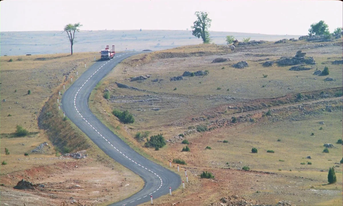 Betty Blue (1986) movie still: wide — A winding asphalt road curves through a dry, sparse, and arid highland terrain. A truck drives along the road…