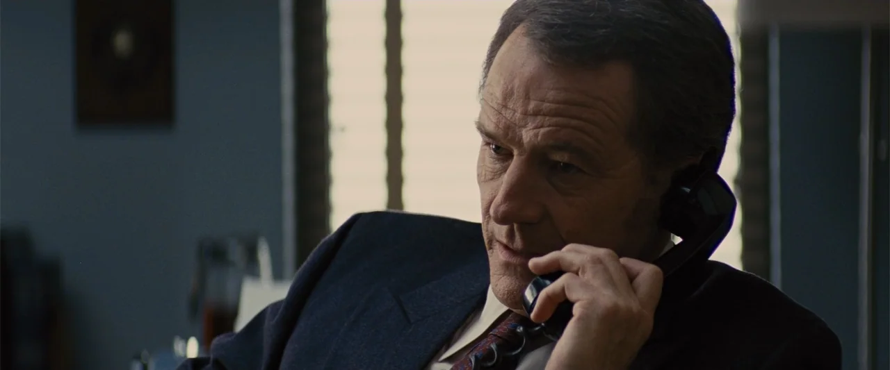 Argo (2012) movie still: medium — An interior office space features horizontal window blinds filtering light against a blue wall. A middle-aged man…