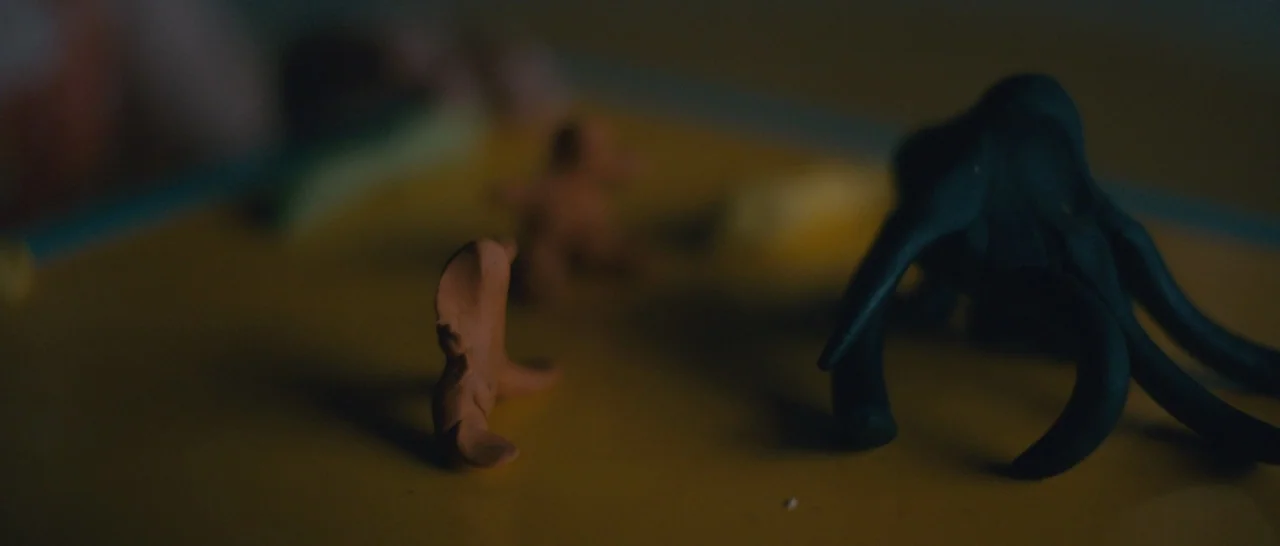 Arrival (2016) movie still: closeup — A dark, out-of-focus environment features a yellow surface holding small, hand-modeled clay sculptures. A…