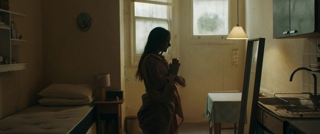 Saint Maud (2019) movie still: medium — A small studio apartment features a bed, a dining table, and a kitchenette area. A young woman stands in the…