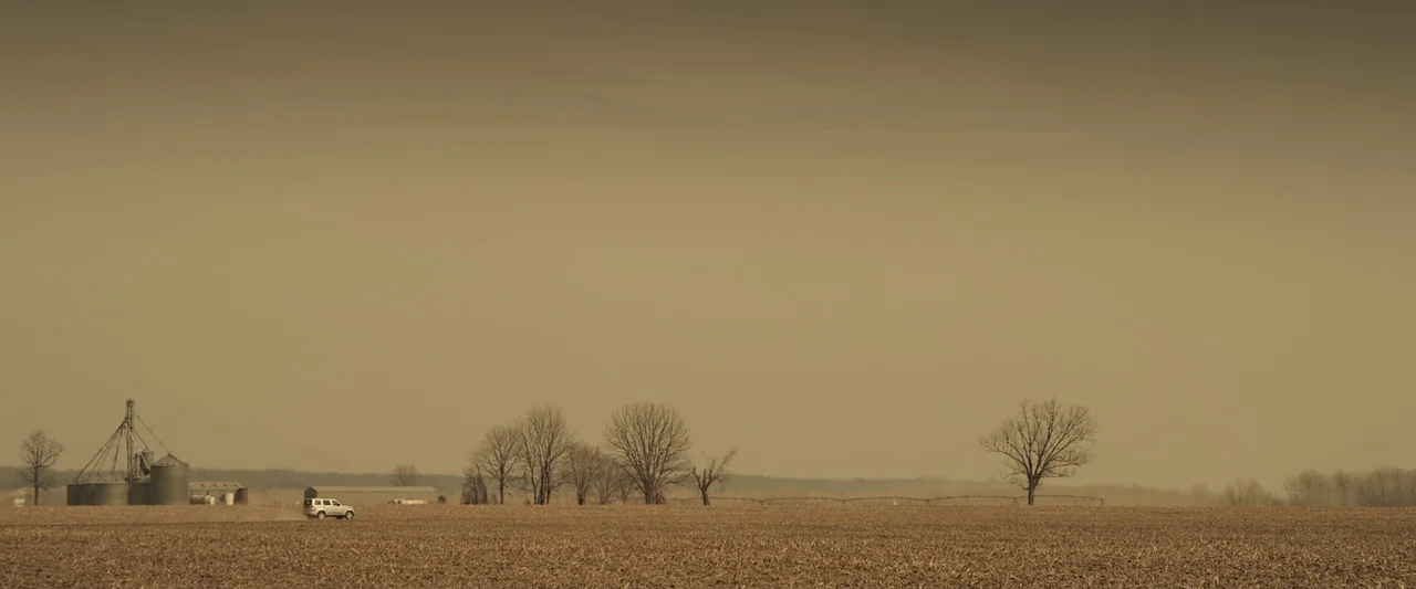 You’re Next (2013) movie still: wide — A vast, barren field stretches toward a distant grain silo and leafless trees. A single white vehicle moves…